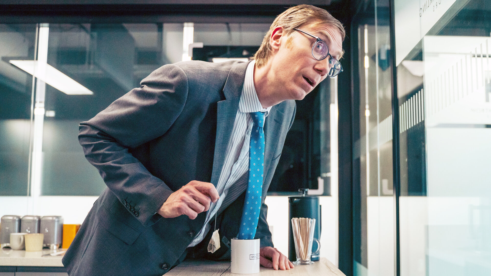 "The Outlaws - Geschäft ist Geschäft": Greg (Stephen Merchant) beugt sich im Büro über den Tisch. Dabei hängt seine Krawatte in einer Kaffeetasse.