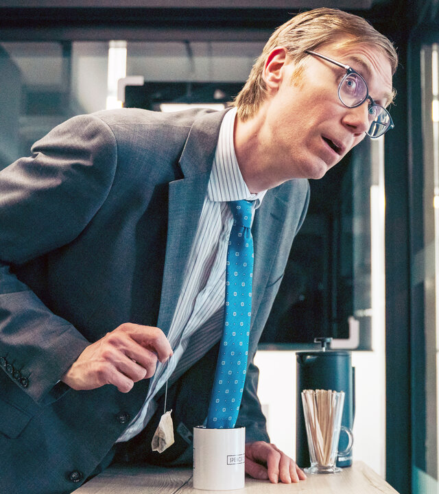 "The Outlaws - Geschäft ist Geschäft": Greg (Stephen Merchant) beugt sich im Büro über den Tisch. Dabei hängt seine Krawatte in einer Kaffeetasse.