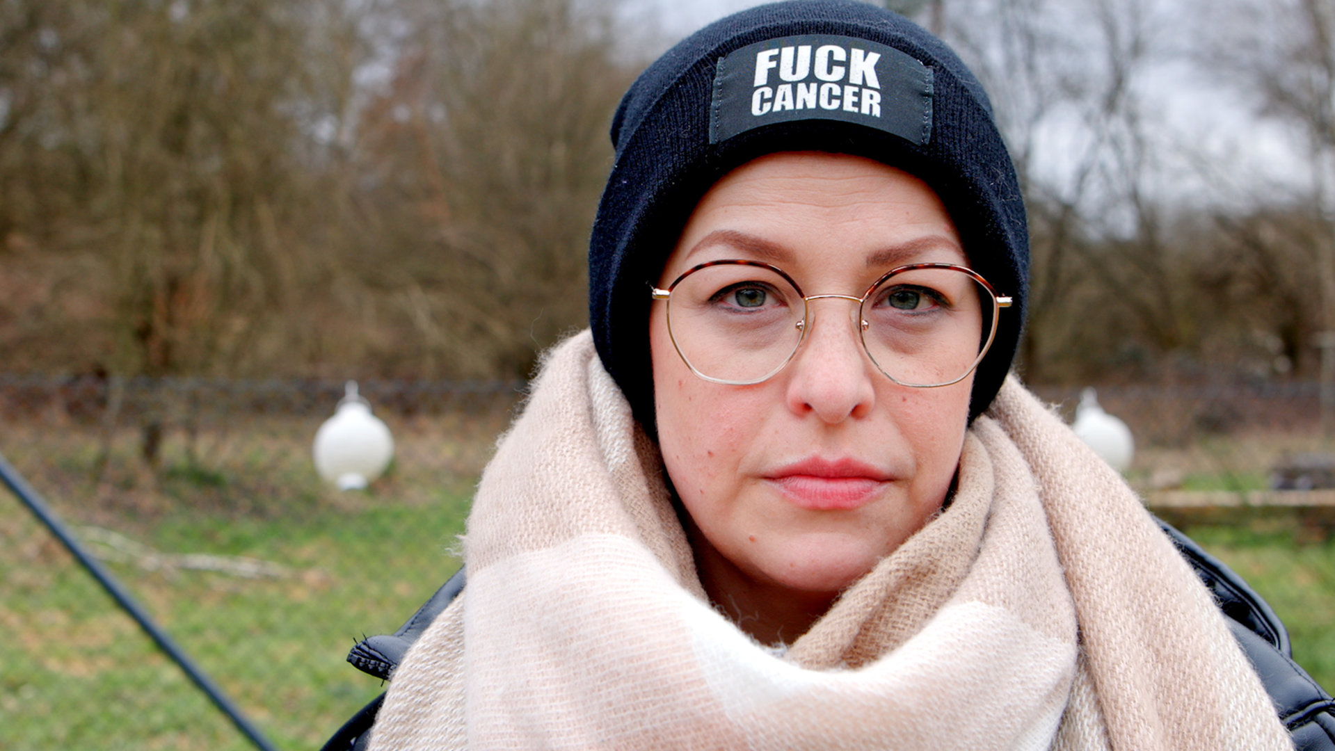 "Mein Kampf gegen Brustkrebs": Porträtfoto von Charlotte. Sie trägt einen hellen Wollschal, eine dunkelblaue Mütze mit der Aufschrift "Fuck Cancer".