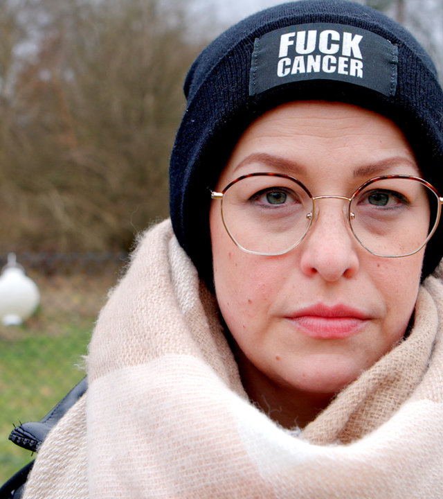 "Mein Kampf gegen Brustkrebs": Porträtfoto von Charlotte. Sie trägt einen hellen Wollschal, eine dunkelblaue Mütze mit der Aufschrift "Fuck Cancer".