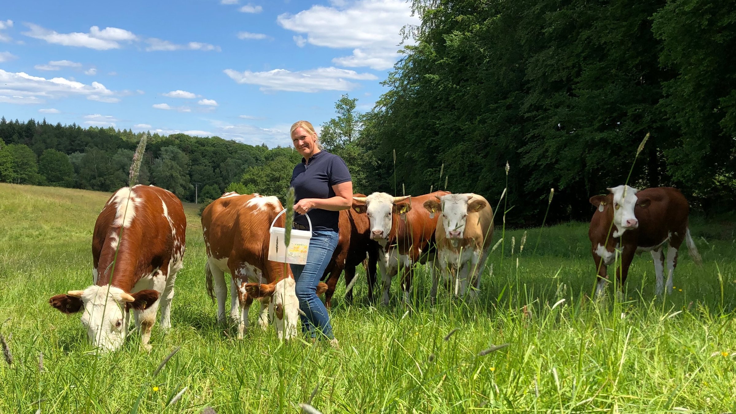 "Terra Xpress - Besser essen und besser leben - Landwirte im Stress": Dana Schwarz steht auf der Grasweide mit einem Eimer in der Hand und schaut in die Kamera. Um sie herum stehen sechs ihrer Kühe.