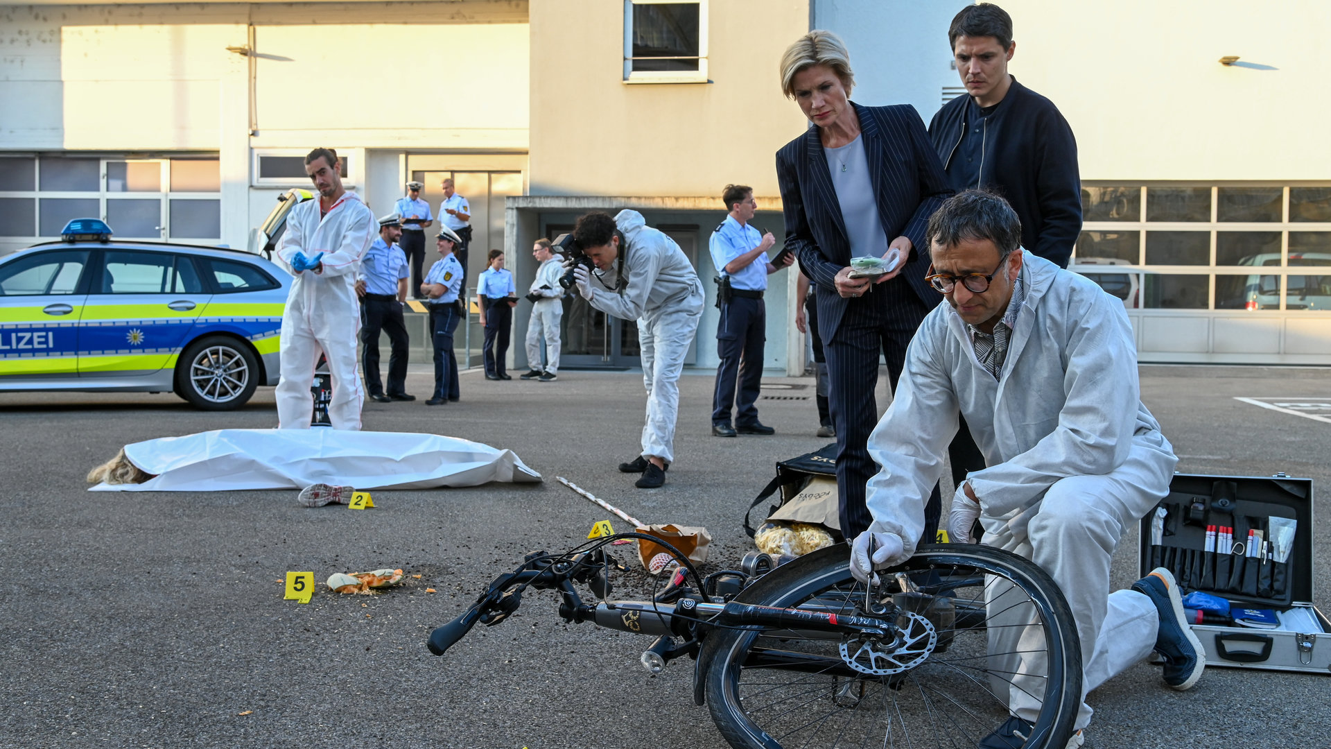 "SOKO Stuttgart - Bikefoodys": Jan Arnaud (Mike Zaka Sommerfeldt) untersucht auf dem Parkplatz vor einem Industriegebäude das verbogene, schwarze Fahrrad des Opfers. Hinter ihm stehen Kriminalhauptkommissarin Martina Seiffert (Astrid M. Fünderich) und IT-Spezialist Rico Sander (Benjamin Strecker) und schauen dem Leiter der KTU über die Schulter. Im Hintergrund begutachtet Rechtsmediziner Dr. Benedikt Förster das am Boden liegende Opfer, das mit einem weißen Leichentuch abgedeckt ist. Spurensicherer und Streifenbeamte sind ebenfalls am Tatort und besprechen sich.