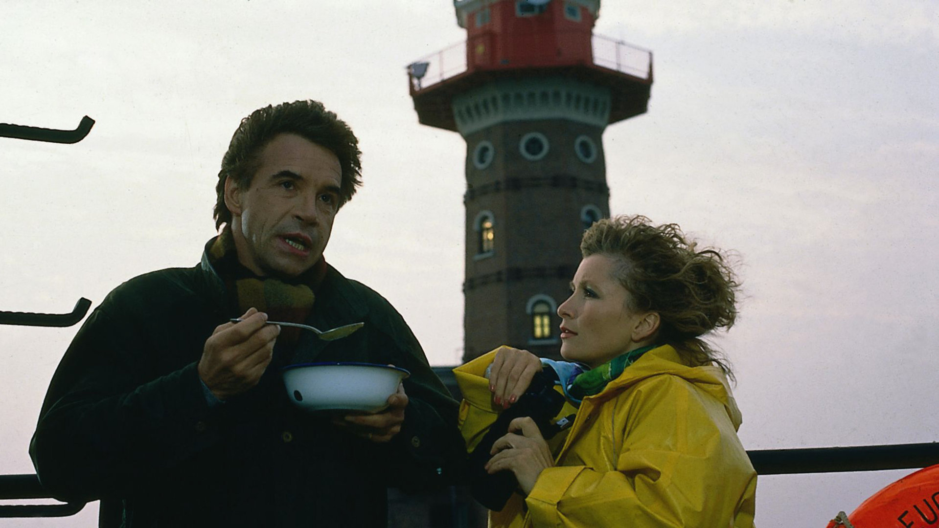 "Das Erbe der Guldenburgs - Die schlechte Freundin": Thomas (Wolf Roth) und Kitty (Susanne Uhlen) stehen vor einem Leuchtturm. Thomas löffelt eine Suppe, Kitty trägt einen gelben Regenmantel.