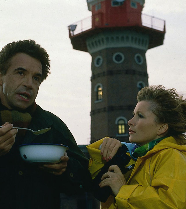 "Das Erbe der Guldenburgs - Die schlechte Freundin": Thomas (Wolf Roth) und Kitty (Susanne Uhlen) stehen vor einem Leuchtturm. Thomas löffelt eine Suppe, Kitty trägt einen gelben Regenmantel.