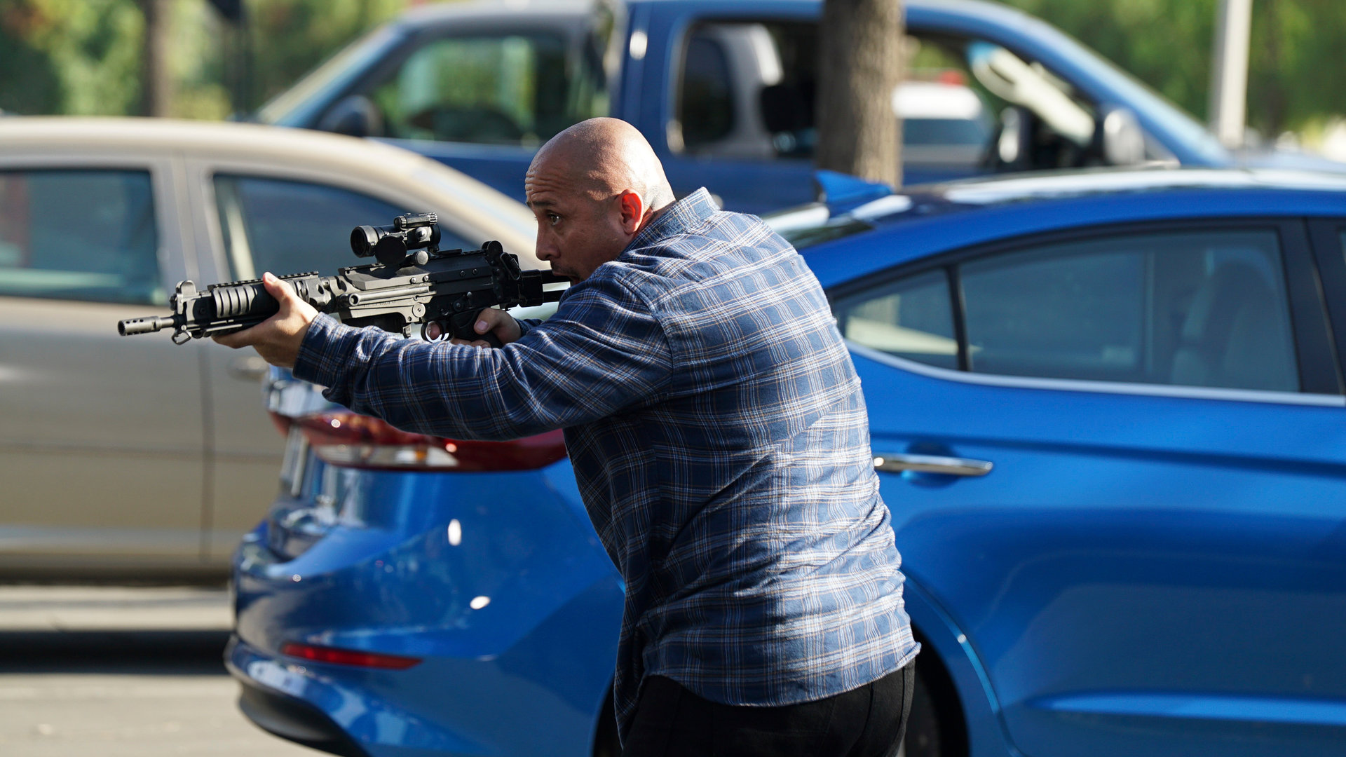 "The Rookie - Der Tausch": Außenaufnahme auf einem Parkplatz. Ein kahlköpfiger Mann hält mit beiden Händen ein Gewehr abschussbereit nach vorne.