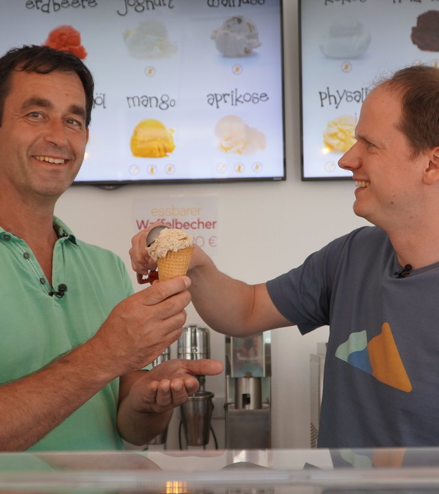 "sonntags - Summer in the City": "sonntags"-Moderator Michael Sahr steht vor einer Schautafel mit verschiedenen Eissorten, hält eine Eiswaffel in der Hand und lächelt Richtung Kamera. Rechts neben ihm steht Eismacher Christoph Heisig, der mit einem Portionierlöffel eine Eiskugel auf die Eiswaffel drückt und Herrn Sahr dabei anschaut.