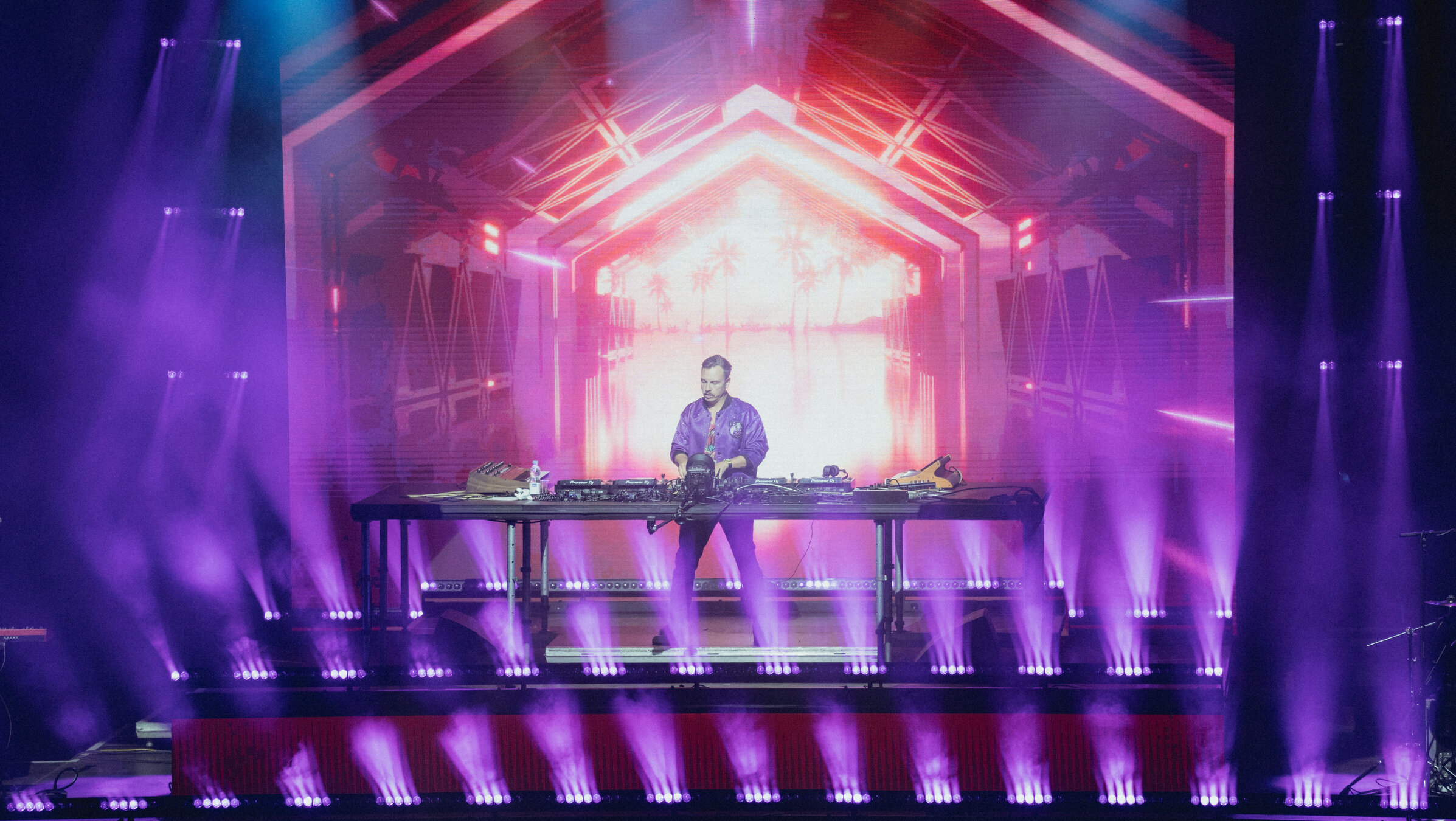 "Purple Disco Machine": Der DJ Purple Disco Machine steht auf der Bühne vor seiner Anlage. Im Hintergrund Lichtinstallationen in blauen, lila und pinken Farbtönen.