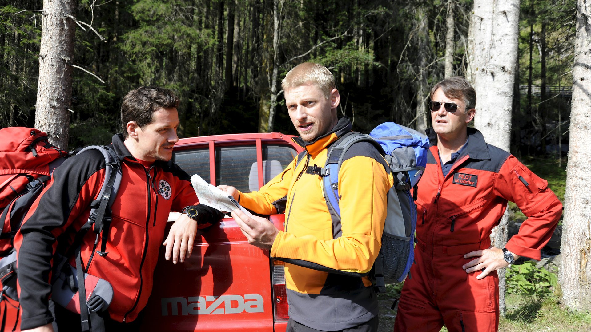"Die Bergretter - Mit dem Rücken zur Wand (2)": Andreas (Martin Gruber) und seine Kollegen Tobias (Markus Brandl) und Michael (Robert Lohr) stehen einsatzbereit mit Ausrüstung am Waldrand. Andreas hält eine Karte in der Hand und schaut in eine Richtung außerhalb des Bildes.