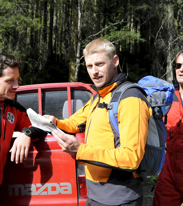 "Die Bergretter - Mit dem Rücken zur Wand (2)": Andreas (Martin Gruber) und seine Kollegen Tobias (Markus Brandl) und Michael (Robert Lohr) stehen einsatzbereit mit Ausrüstung am Waldrand. Andreas hält eine Karte in der Hand und schaut in eine Richtung außerhalb des Bildes.