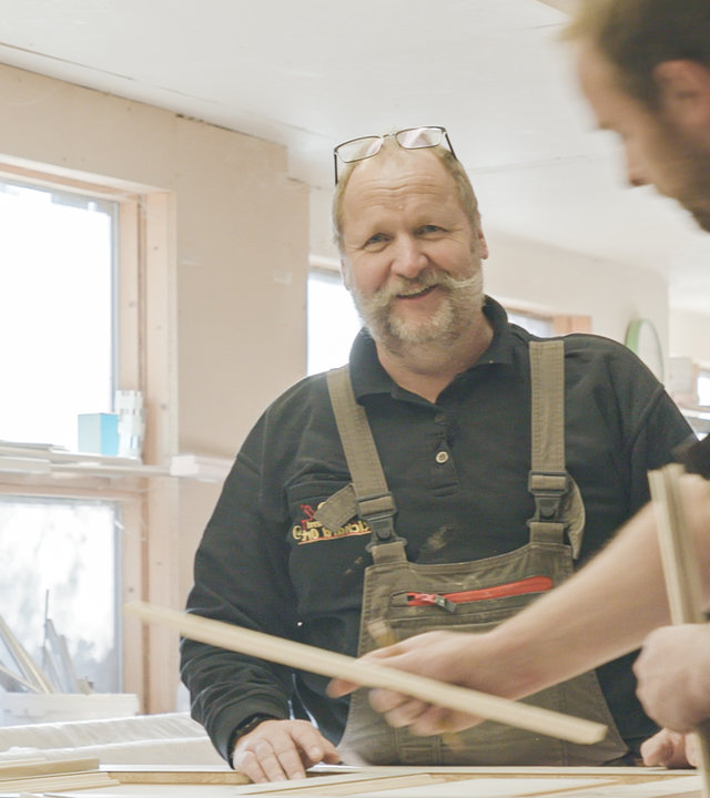 "37° Jetzt bestimme ich! - Generationswechsel im Familienbetrieb": Ein Mann mit grauen Haaren und einem Schnurrbart schaut lächelnd in die Kamera. Er trägt eine Arbeitshose und ein hält ein Stück Holz in der Hand. Im Vordergrund verschwommen ein junger Mann, der ebenfalls ein Stück Holz in jeder Hand hält.