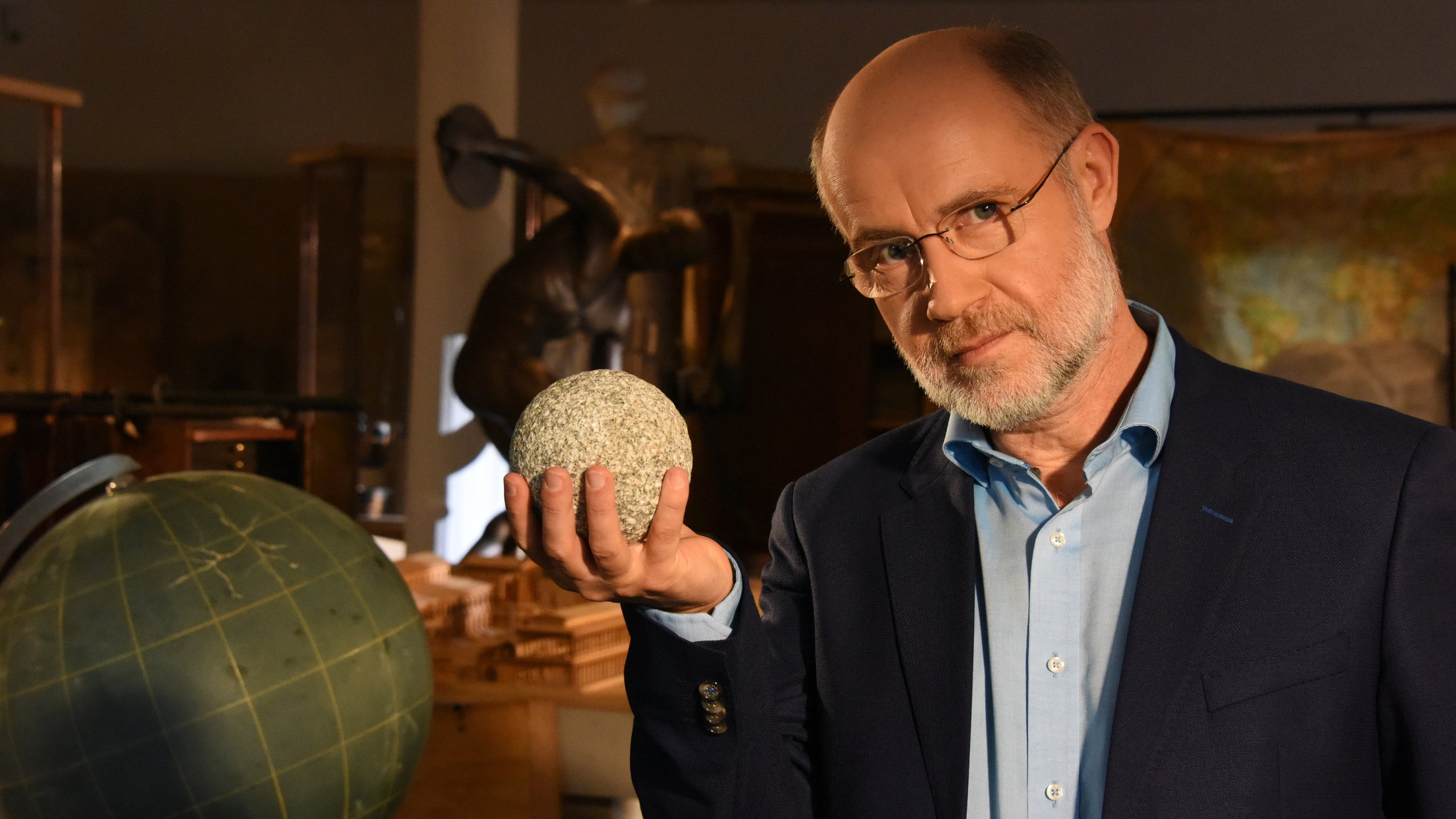 "Terra X: Ungelöste Fälle der Archäologie: Verlorenes Wissen": Harald Lesch steht mit einer kleinen Steinkugel in der Hand inmitten von altertümlichen Requisiten.