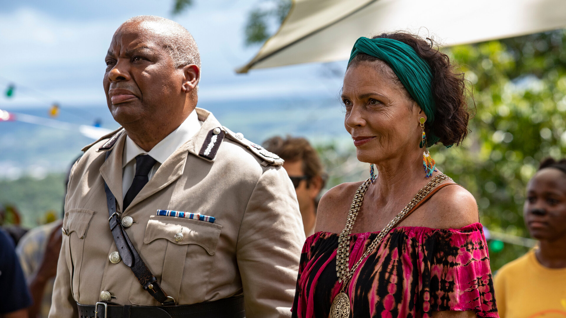 "Death in Paradise - Der Rückkehrer": Commissioner Selwyn Patterson (Don Warrington) und Catherine Bordey (Elizabeth Bourgine) stehen zusammen mit anderen Personen draußen und blicken in Richtung Bühne.