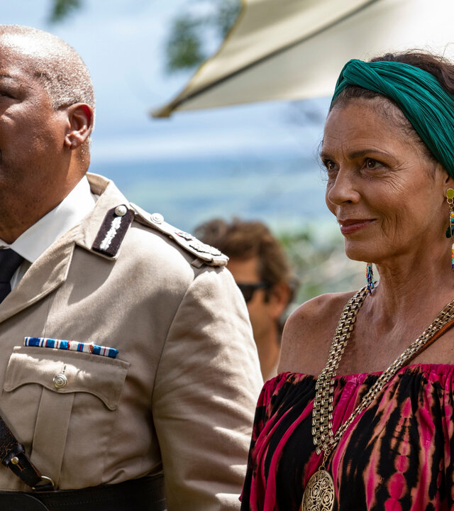 "Death in Paradise - Der Rückkehrer": Commissioner Selwyn Patterson (Don Warrington) und Catherine Bordey (Elizabeth Bourgine) stehen zusammen mit anderen Personen draußen und blicken in Richtung Bühne.