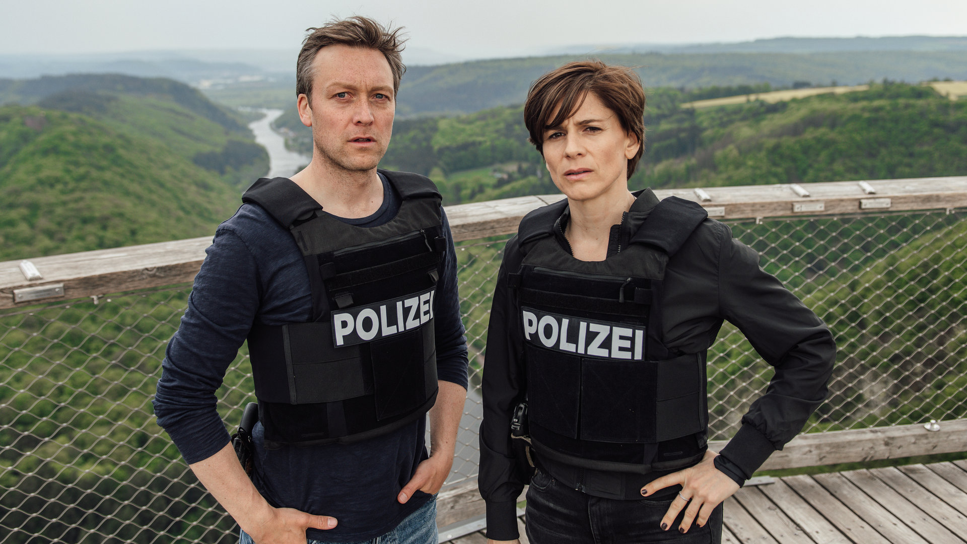 „In Wahrheit – Blind vor Liebe“: Das Ermittlerteam Judith Mohn (Christina Hecke) und Freddy Breyer (Robin Sondermann) steht auf einer Aussichtsplattform mit Ausblick auf die Saarschleife. Sie tragen beide Schutzwesten.