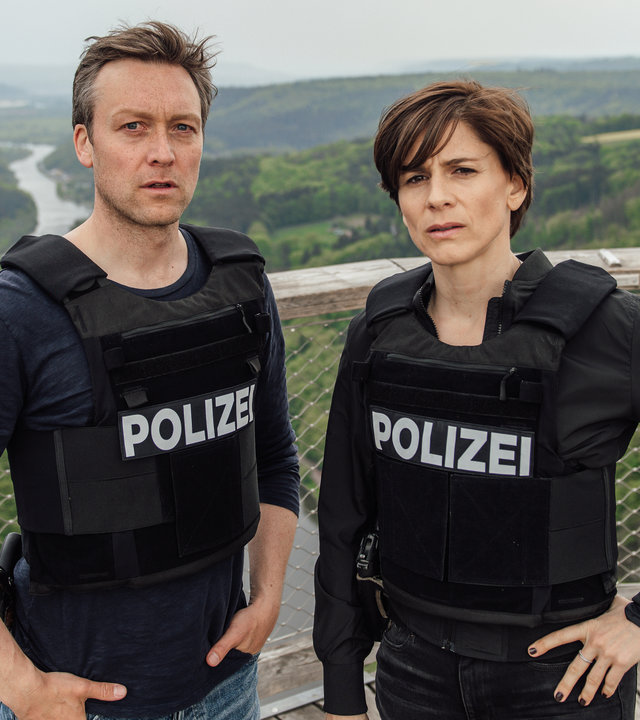 „In Wahrheit – Blind vor Liebe“: Das Ermittlerteam Judith Mohn (Christina Hecke) und Freddy Breyer (Robin Sondermann) steht auf einer Aussichtsplattform mit Ausblick auf die Saarschleife. Sie tragen beide Schutzwesten.