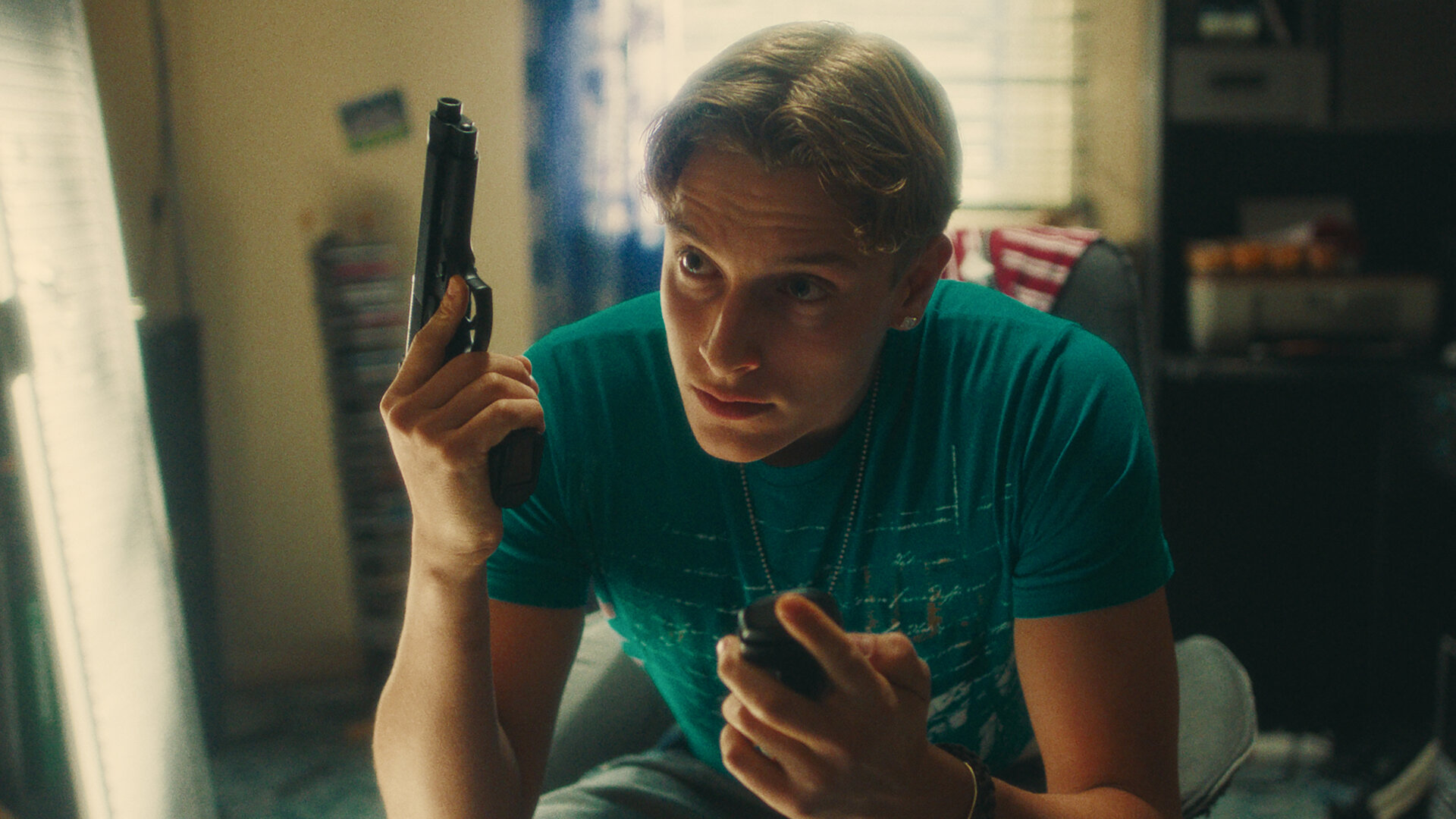 "Chabos - Ocean's Four": PD (Jonathan Kriener) sitzt in einem Jugendzimmer. In der linken Hand hat er sein Klapphandy, in der rechten eine Softair-Pistole.