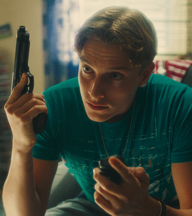 "Chabos - Ocean's Four": PD (Jonathan Kriener) sitzt in einem Jugendzimmer. In der linken Hand hat er sein Klapphandy, in der rechten eine Softair-Pistole.