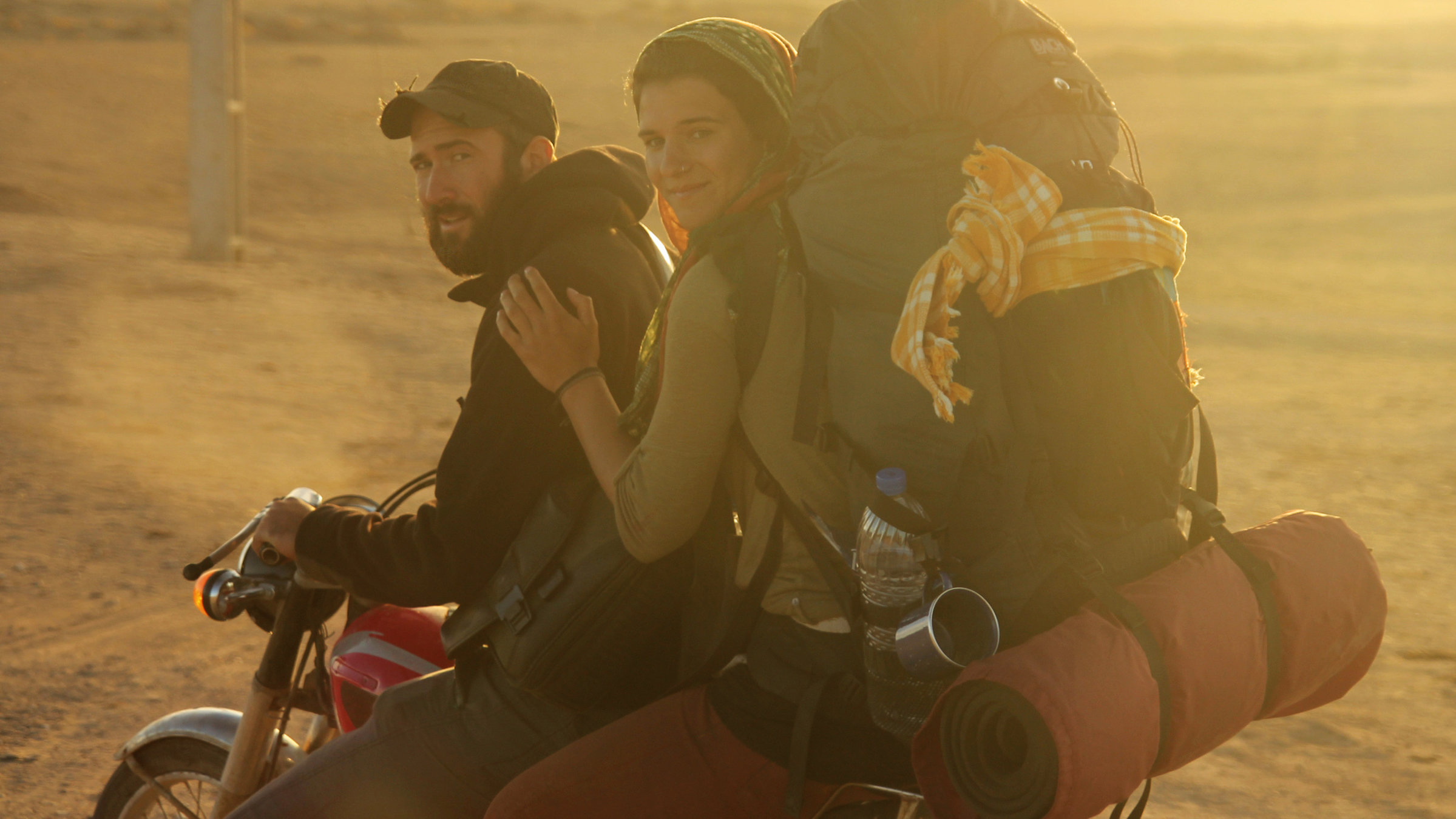 „Weit“: Ein junger Mann mit Bart und Baseballkappe sitzt am Lenker eines Motorrads und schaut in die Kamera. Hinter ihm sitzt eine junge Frau, die sich an seiner Schulter festhält, die Haare mit einem Tuch bedeckt hat und einen vollbepackten Rucksack auf den Schultern trägt. Im Hintergrund ist eine weite Steppenlandschaft unscharf und in einem goldgelben Licht der Abendsonne zu sehen.