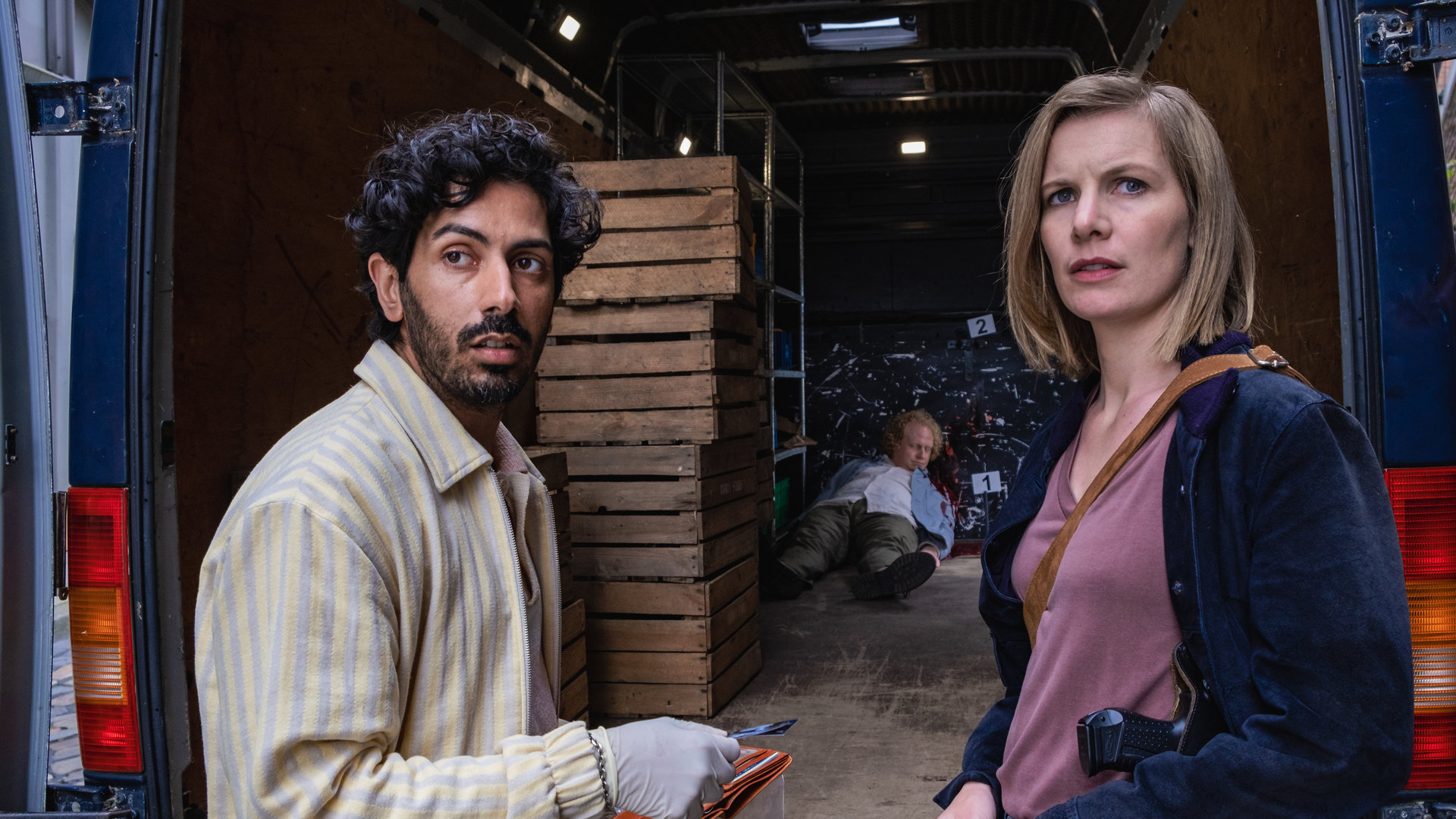 "SOKO Hamburg - Marktschreie": Luiz Vieira dos Santos (Thiago Braga de Oliveira) und Lena Testorp (Anna von Haebler) stehen nebeneinander vor der geöffneten Tür eines Lieferwagens und blicken ernst nach vorne. Luiz hält eine Brieftasche in der Hand, aus der er eine Karte zieht. Er trägt weiße Gummihandschuhe. Aus Lenas dunkelblauer Jacke ragt der Griff ihrer Pistole. Im Lagerraum sieht man braune Kisten, die übereinander gestapelt wurden. Im hinteren Teil liegt der tote Sven Peters (Julian Greis) auf dem Rücken.