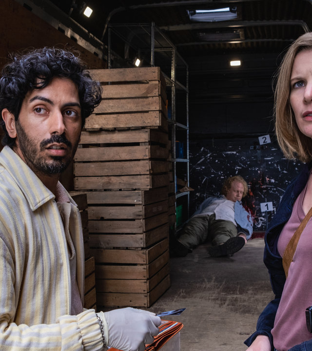 "SOKO Hamburg - Marktschreie": Luiz Vieira dos Santos (Thiago Braga de Oliveira) und Lena Testorp (Anna von Haebler) stehen nebeneinander vor der geöffneten Tür eines Lieferwagens und blicken ernst nach vorne. Luiz hält eine Brieftasche in der Hand, aus der er eine Karte zieht. Er trägt weiße Gummihandschuhe. Aus Lenas dunkelblauer Jacke ragt der Griff ihrer Pistole. Im Lagerraum sieht man braune Kisten, die übereinander gestapelt wurden. Im hinteren Teil liegt der tote Sven Peters (Julian Greis) auf dem Rücken.