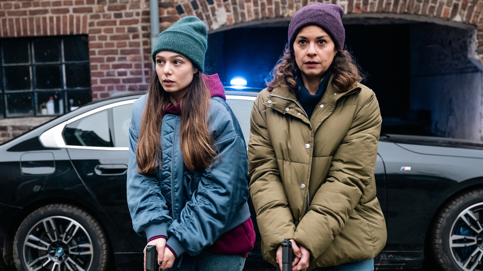 SOKO Köln - Jagdzeit": Amira Mahdi (Aylin Ravanyar) und Lilly Funke (Tatjana Kästel) stehen nebeneinander vor einem schwarzen Auto und halten ihre gezückten Waffen nach unten gerichtet vor den Körper. Amira sieht aufmerksam zur Seite, während Lilly angespannt nach vorne blickt.
