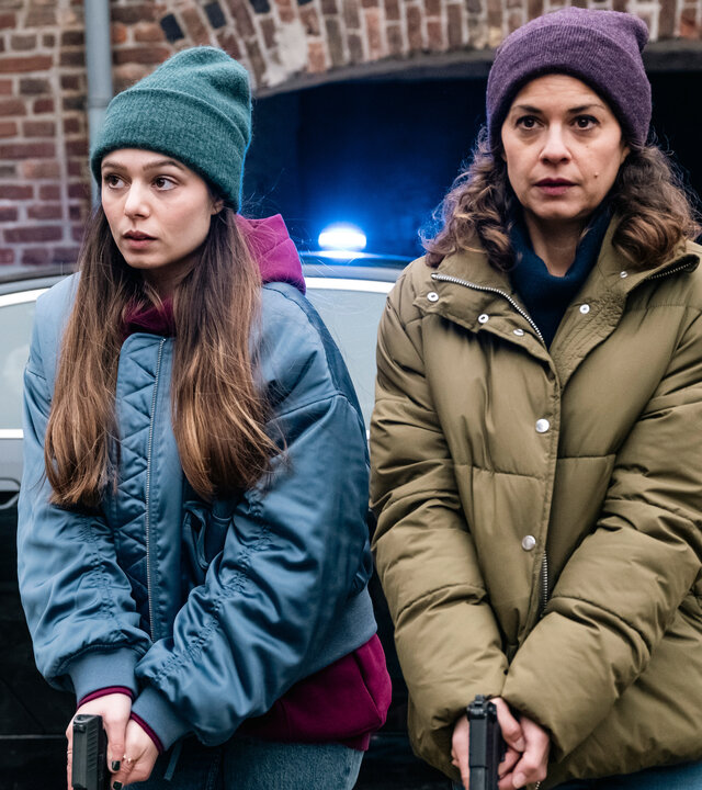 SOKO Köln - Jagdzeit": Amira Mahdi (Aylin Ravanyar) und Lilly Funke (Tatjana Kästel) stehen nebeneinander vor einem schwarzen Auto und halten ihre gezückten Waffen nach unten gerichtet vor den Körper. Amira sieht aufmerksam zur Seite, während Lilly angespannt nach vorne blickt.