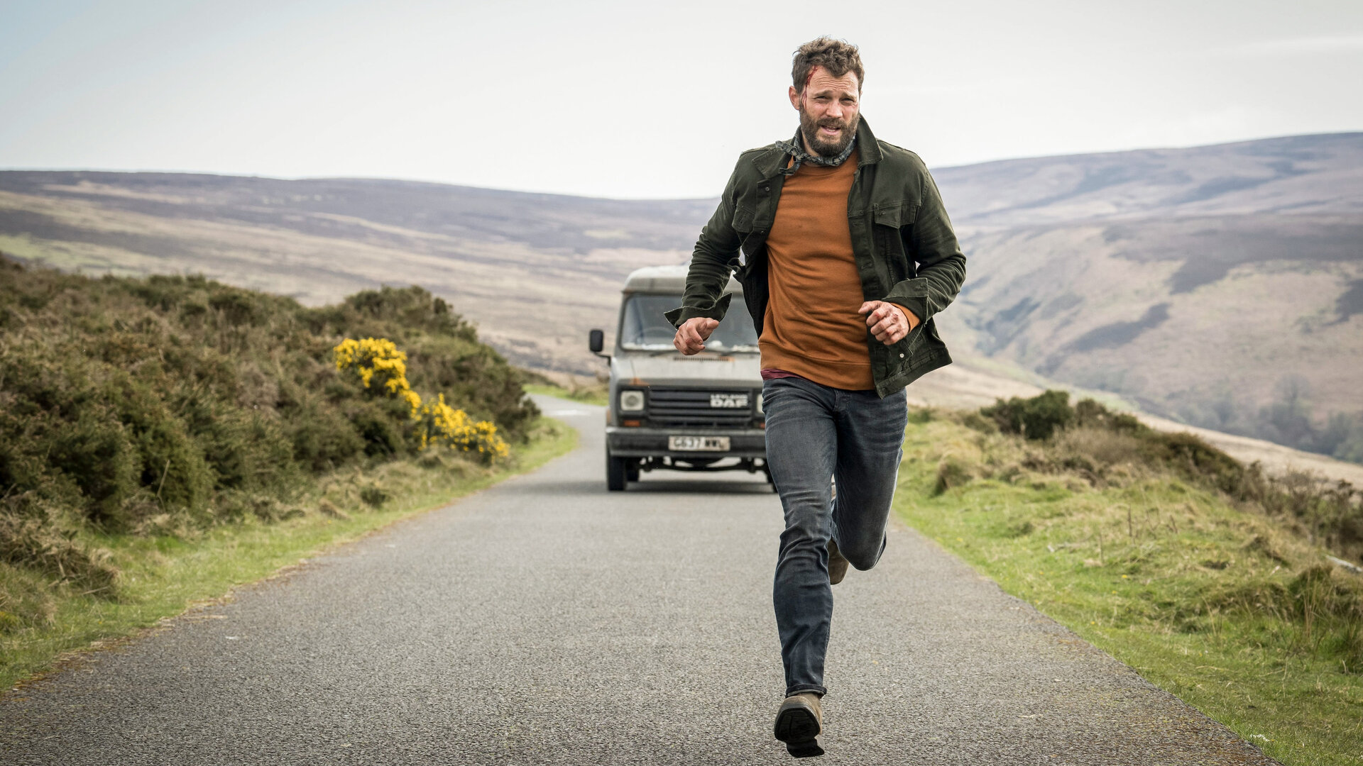 "The Tourist - Irisches Blut - Iren sind menschlich": Elliot Stanley (Jamie Dornan), gekleidet in Jeans, einem rotockerfarbenen Shirt, einem Halstuch und einer leichten Jacke, läuft auf einer Landstraße in einer irischen Hügellandschaft vor einem olivfarbenen Kleintransporter davon. An seiner Stirn blutet eine Platzwunde.