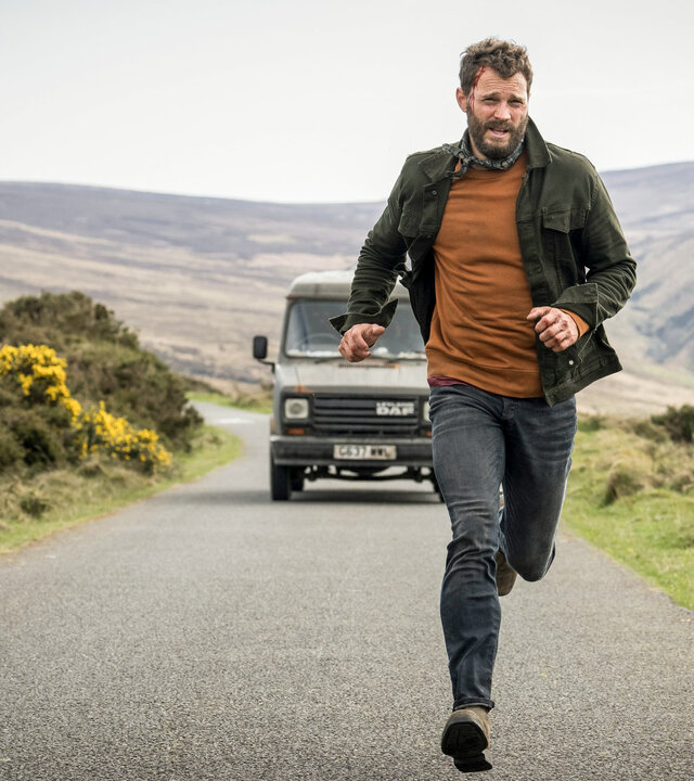 "The Tourist - Irisches Blut - Iren sind menschlich": Elliot Stanley (Jamie Dornan), gekleidet in Jeans, einem rotockerfarbenen Shirt, einem Halstuch und einer leichten Jacke, läuft auf einer Landstraße in einer irischen Hügellandschaft vor einem olivfarbenen Kleintransporter davon. An seiner Stirn blutet eine Platzwunde.