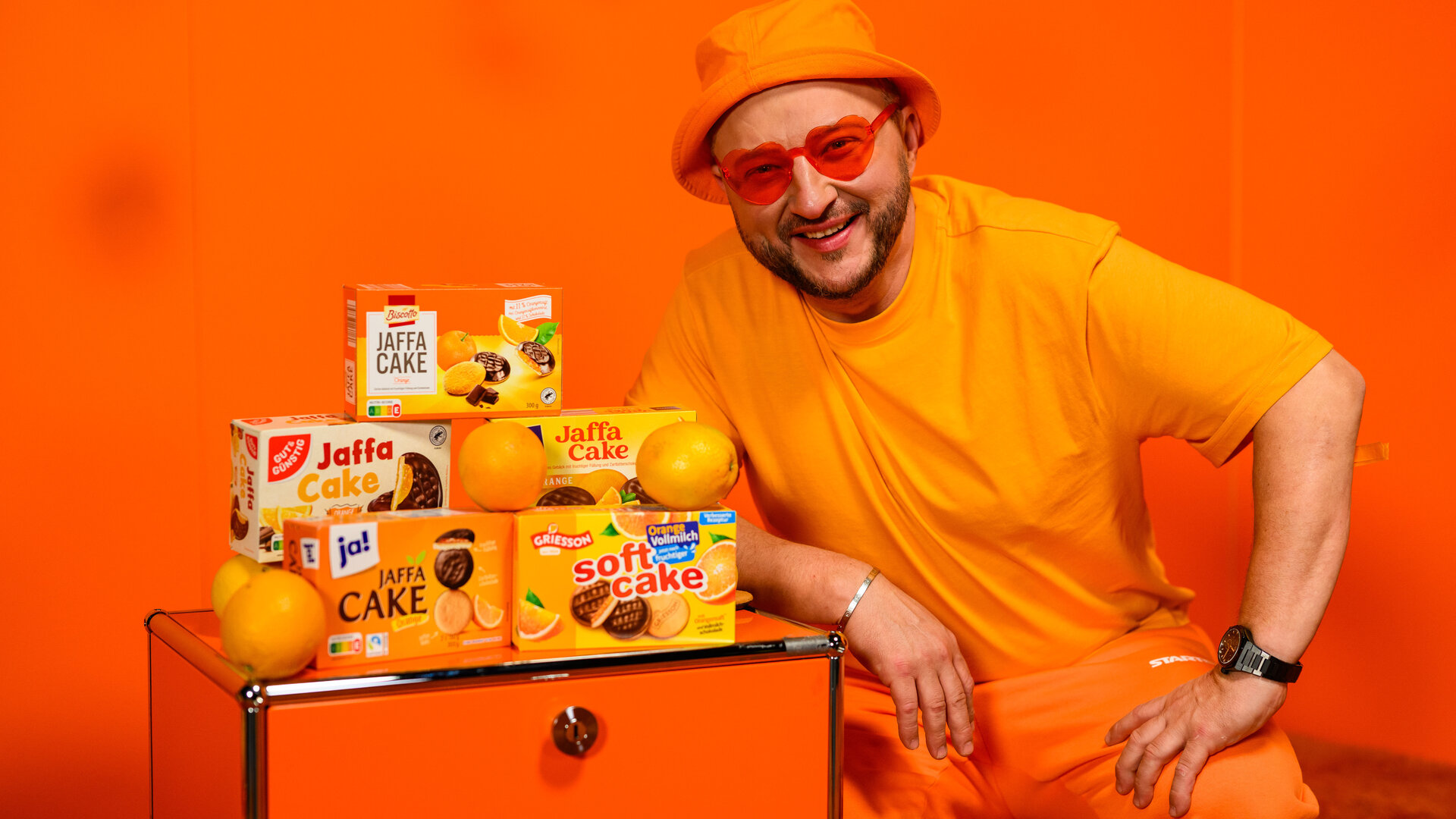 "besseresser: Die Tricks in Bärchenwurst, Pizzateig & Co.": Sebastian Lege steht vor orangefarbenem Hintergrund neben einer orangefarbenen Kommode, auf der Jaffa Cake Produkte verschiedener Marken stehen.