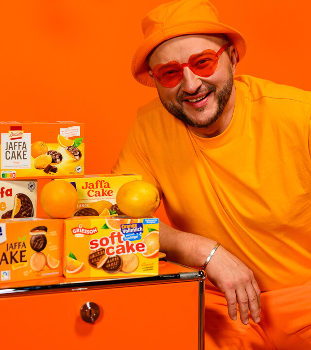 "besseresser: Die Tricks in Bärchenwurst, Pizzateig & Co.": Sebastian Lege steht vor orangefarbenem Hintergrund neben einer orangefarbenen Kommode, auf der Jaffa Cake Produkte verschiedener Marken stehen.