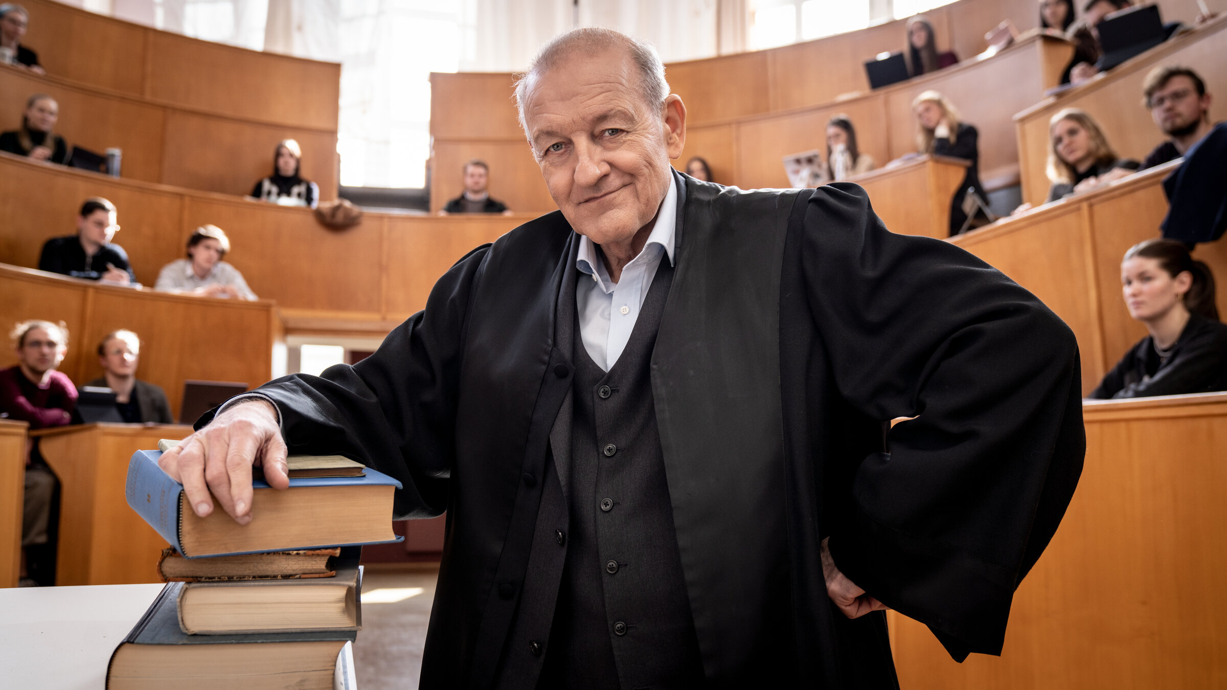 "Wilsberg - Über dem Gesetz": Wilsberg (Leonard Lansink) steht in einem Unihörsaal, sein Arm lehnt auf einem Stapel mit dicken Büchern, er blickt lächelnd in die Kamera. In den Sitzreihen hinter ihm sitzen Studierende und schauen in Richtung Wilsberg.