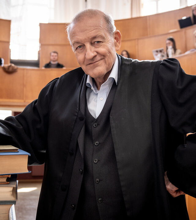 "Wilsberg - Über dem Gesetz": Wilsberg (Leonard Lansink) steht in einem Unihörsaal, sein Arm lehnt auf einem Stapel mit dicken Büchern, er blickt lächelnd in die Kamera. In den Sitzreihen hinter ihm sitzen Studierende und schauen in Richtung Wilsberg.