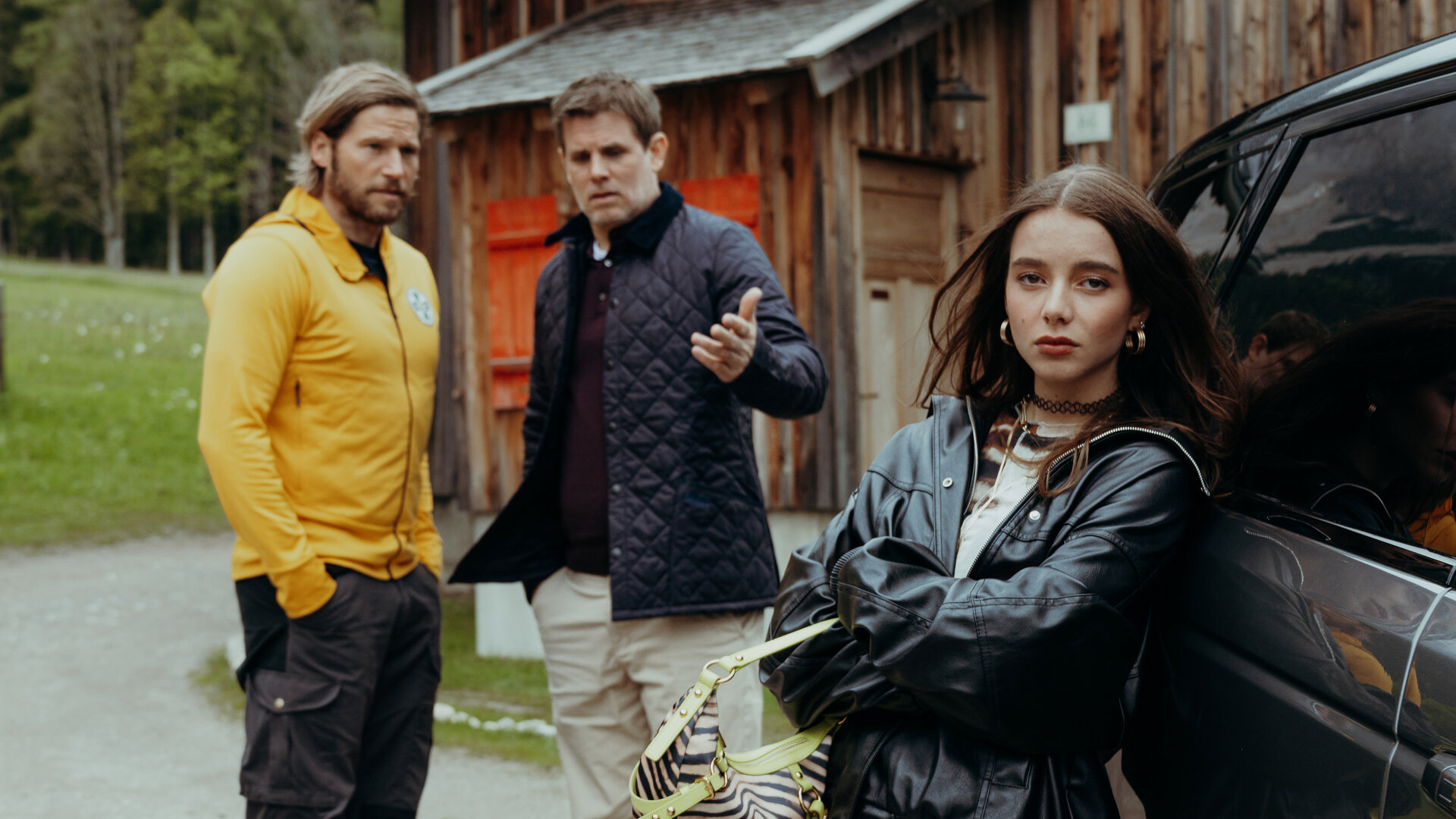 "Die Bergretter: Vertrauen": Auf Emilies Hof:  Eine zornig wirkende Mia Steiner (Lisa Junick) lehnt, die Arme vor der Brust verschränkt, an einem Auto und blickt in die Kamera. Im Hintergrund stehen Markus Kofler (Sebastian Ströbel) und Lorenz König (Steffen Groth) und unterhalten sich. Lorenz zeigt dabei mit der Hand auf Mia.