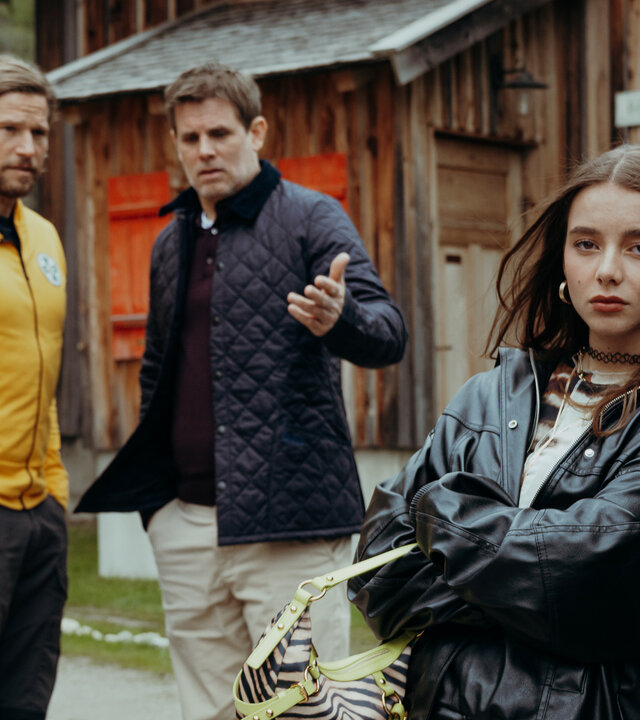 "Die Bergretter: Vertrauen": Auf Emilies Hof:  Eine zornig wirkende Mia Steiner (Lisa Junick) lehnt, die Arme vor der Brust verschränkt, an einem Auto und blickt in die Kamera. Im Hintergrund stehen Markus Kofler (Sebastian Ströbel) und Lorenz König (Steffen Groth) und unterhalten sich. Lorenz zeigt dabei mit der Hand auf Mia.