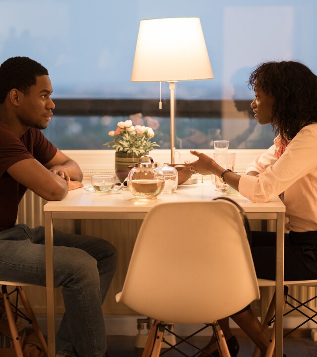 "SOKO Wien - Die Dolmetscherin": Hamza Biobaku (Gabriel-Winner Amorin) und Ayo Ajiri (Edith Saldanha) sitzen sich an einem Tisch vor einem Fenster gegenüber und schauen sich an. Draußen dämmert es. Auf dem Tisch stehen Wassergläser und eine angeschaltete Lampe.
