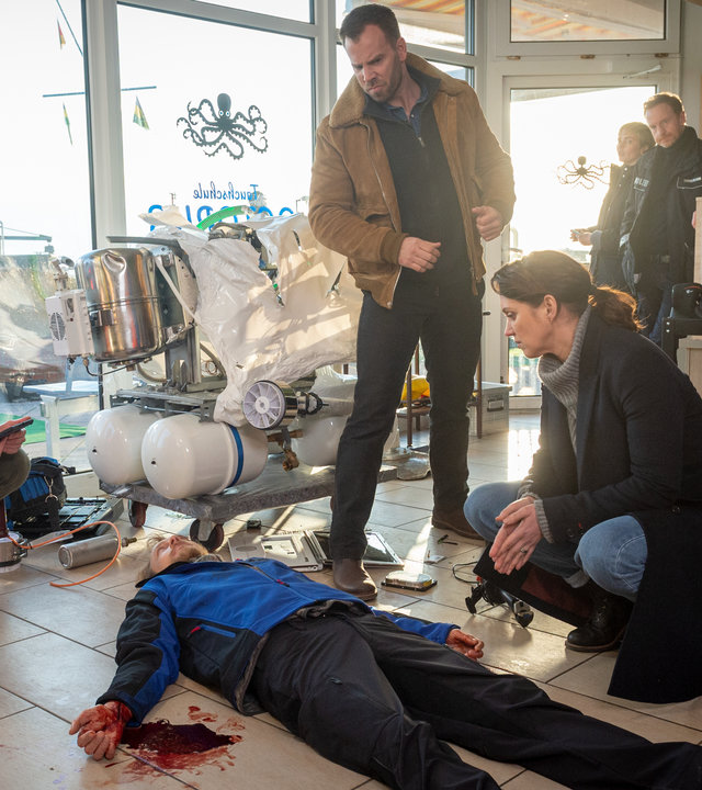 "SOKO Wismar – Geisternetze“: In der Tauchschule liegt Falk Leonhardt (Marcus Kuhn) mit blutigen Händen tot auf dem Boden. Hinter ihm steht ein demoliertes Gerät mit weißen Tanks. Helene Sturbeck (Katharina Blaschke), Pöhlmann (Dominic Boeer) und Joost (Nike Fuhrmann) untersuchen die Leiche, im Hintergrund stehen Stine (Sidsel Hindhede) und Timmermann (Mathias Junge).