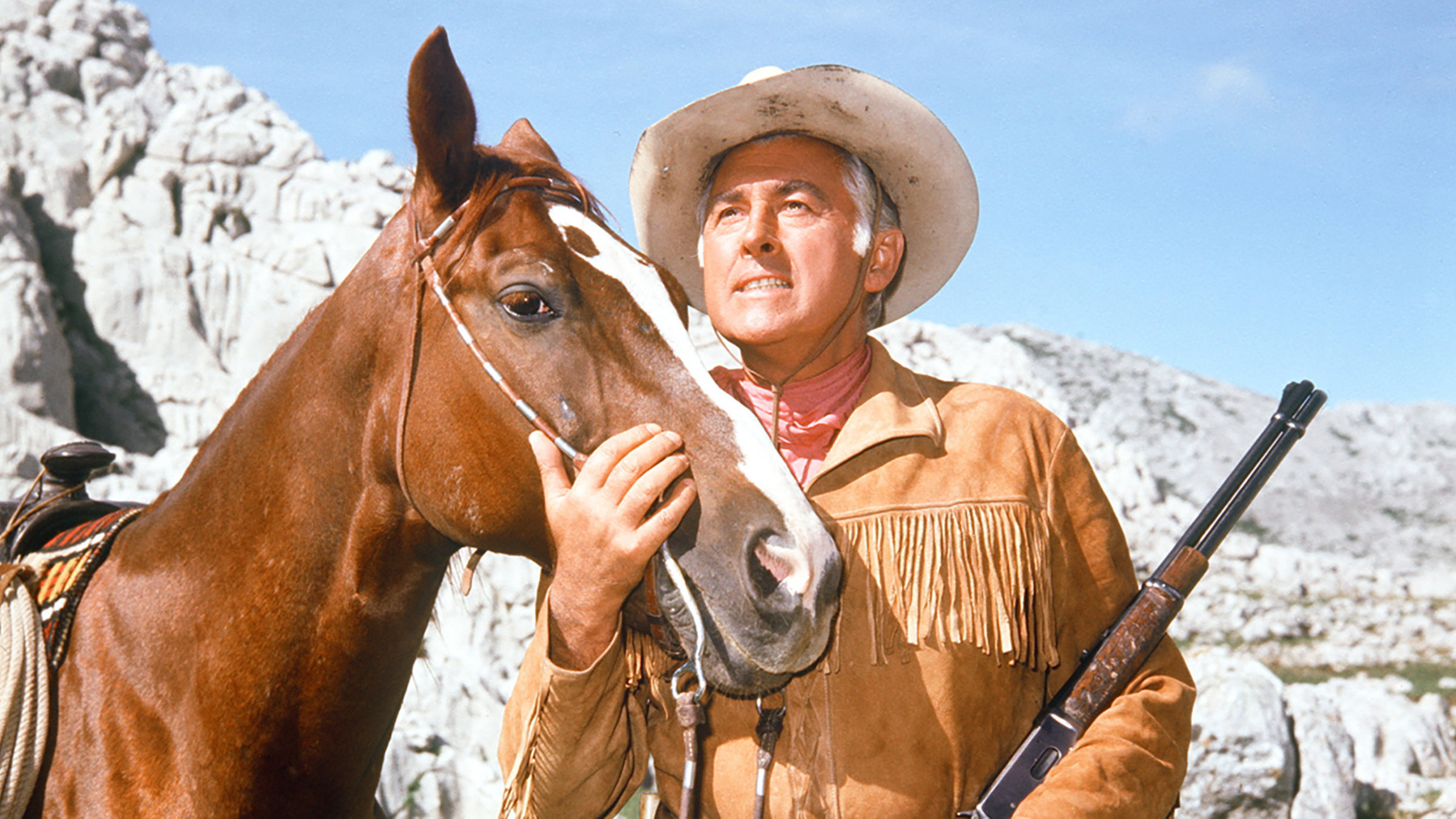 "Old Surehand 1. Teil": Old Surehand (Stewart Granger) in der Nahaufnahme, wie er rechts neben einem Pferd steht. Er hält ein Gewehr in der Hand und schaut nachdenklich in die Ferne.