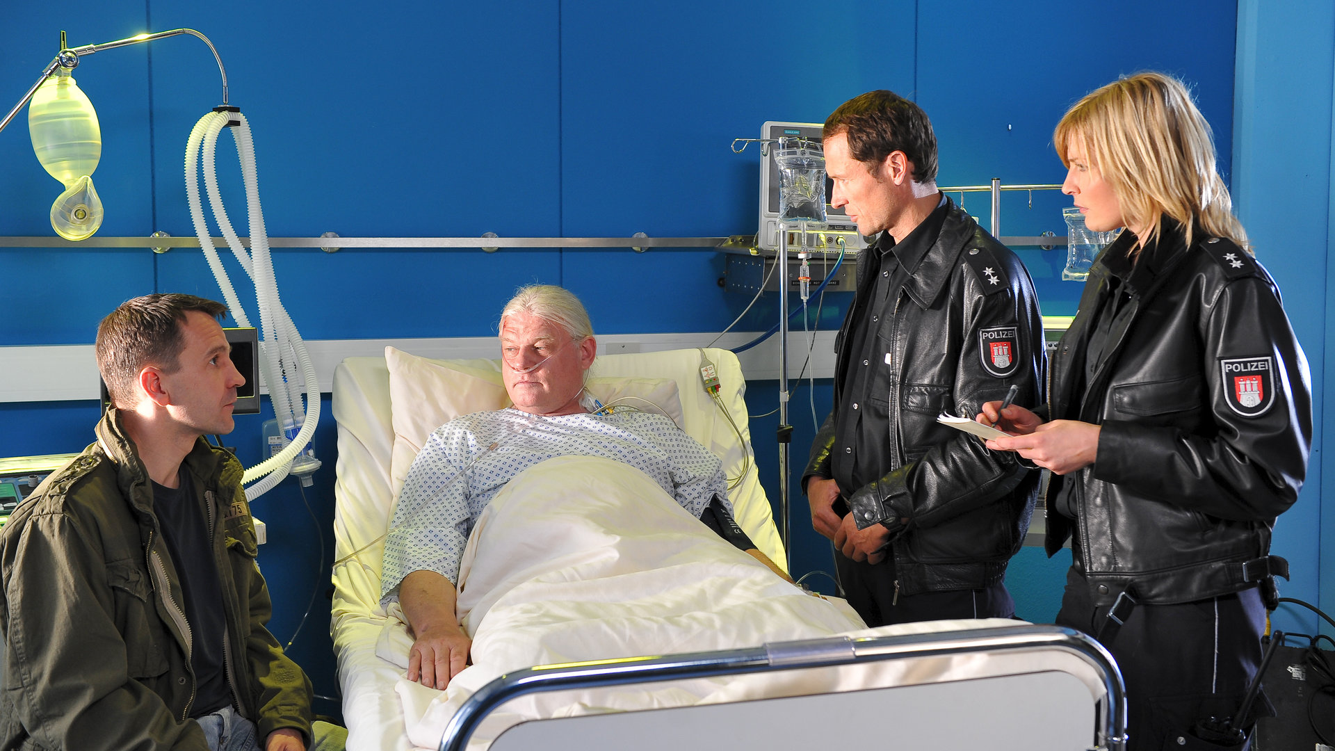 "Notruf Hafenkante - Die große Versuchung": Günther Siebold (Heinrich Rolfing) liegt in einem Krankenhausbett. Neben ihm sitzt sein Kollege Kadde (Michael Ehnert). Melanie (Sanna Englund) und Kai (Markus Knüfken) stehen auf der anderen Seite des Bettes und befragen die beiden.