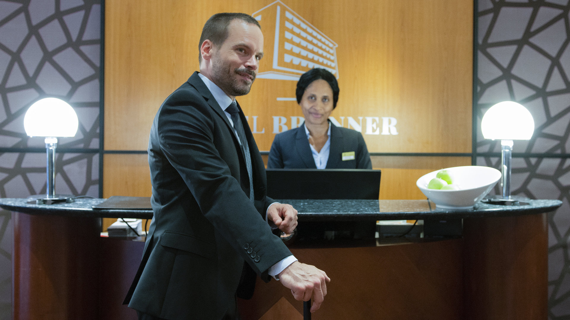 "Ein Fall für zwei - Der Abschiedsbrief": Leo (Wanja Mues) steht im schicken Anzug an der Hotelrezeption. Er stützt sich mit dem linken Arm lässig am Tresen ab und blickt grinsend in die Hotellobby. Eine Rezeptionistin (Komparsin) schaut Leo lächelnd an.