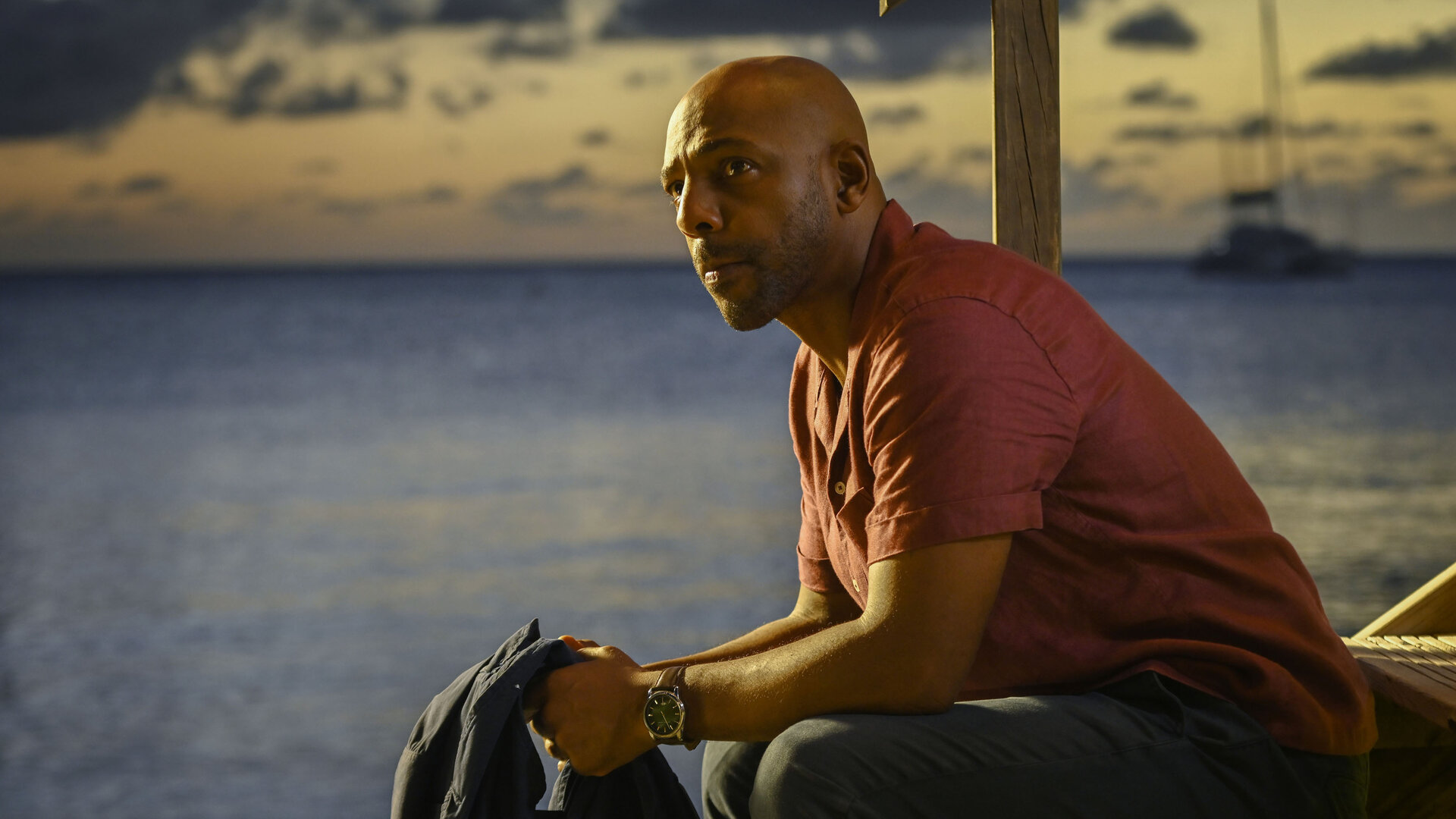 "Death in Paradise - Entscheidungen": Detective Mervin Wilson (Don Gilet) sitzt auf einer hölzernen Treppe vor dem Meer und stützt die Ellenbogen auf den Oberschenkeln ab. Mit fokussiertem Blick schaut er nach vorn.