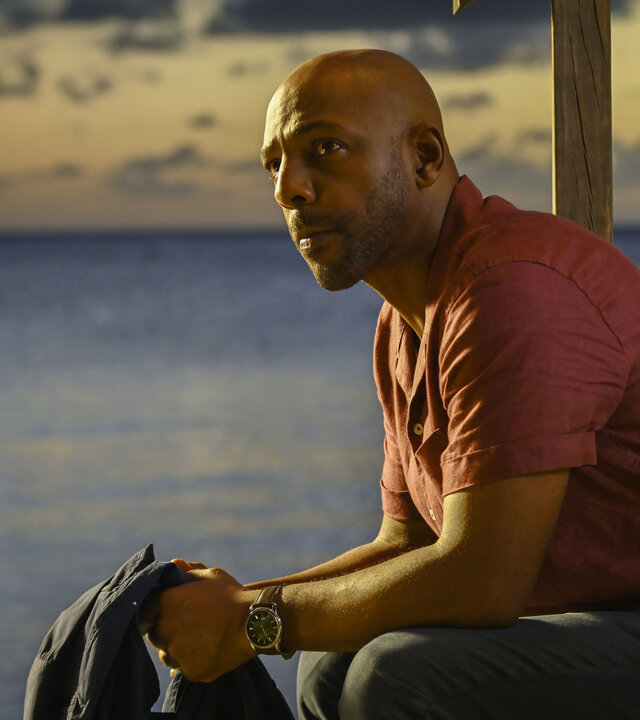 "Death in Paradise - Entscheidungen": Detective Mervin Wilson (Don Gilet) sitzt auf einer hölzernen Treppe vor dem Meer und stützt die Ellenbogen auf den Oberschenkeln ab. Mit fokussiertem Blick schaut er nach vorn.