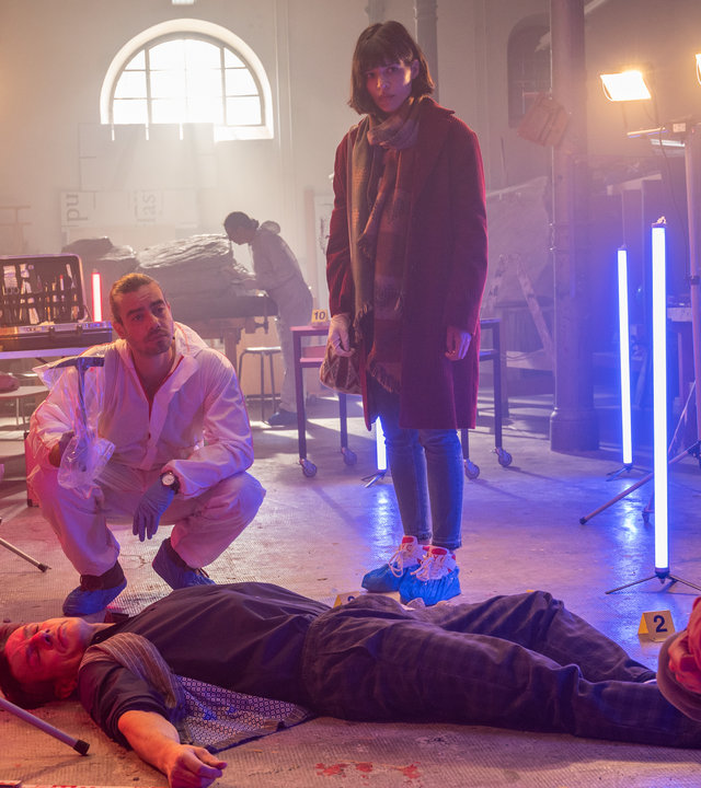 "SOKO Stuttgart – In Stein gemeißelt": Bildhauer und Professor Christian Schilling (Urs Remond) wird erschlagen in einem Atelier der Kunsthochschule gefunden. Kommissarin Lea Gomez (Jasmin Gassmann), ihr Kollege Jo Stoll (Peter Ketnath) und Dr. Benedikt Förster (Florian Wünsche) stehen und knien um den Leichnam herum. Der Rechtsmediziner hält einen eingetüteten Hammer in den Händen. Ein Spurensicherer macht Fotos. Stehleuchten mit Neonlicht färben das Licht des Raumes rot und blau.
