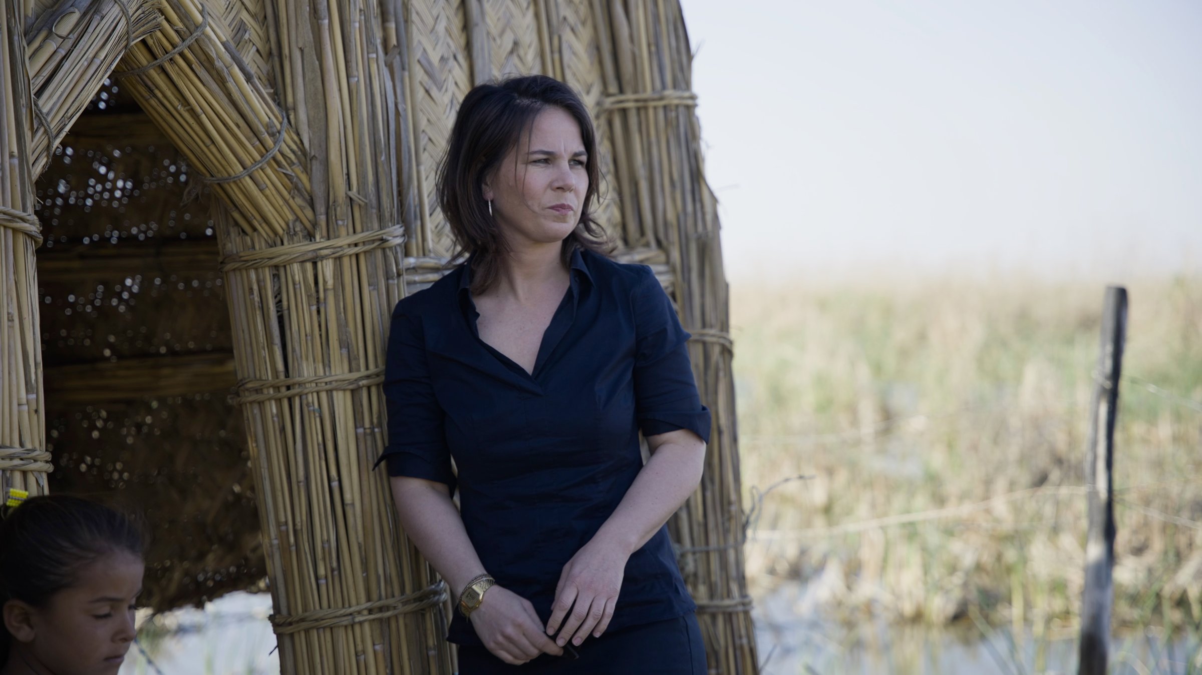 "ZDFzeit: Mensch Baerbock! Die undiplomatische Diplomatin":  Vor einer Hütte aus Bambus steht Ministerin Baerbock, mit beobachtendem Blick.