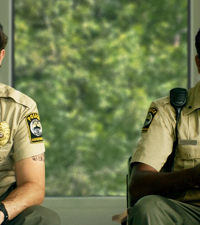 "Greedy People - Gelegenheit macht gierig": Plakatmotiv, Halbnahe: Terry (Joseph Gordon-Levitt) und Will (Himesh Patel) sitzen in Polizeiuniform nebeneinander vor einer Fensterfront mit Blick ins Grüne. Während Terry stoisch nach vorne blickt, zeigt Will einen besorgten Gesichtsausdruck. Beide halten eine Kaffeetasse in der Hand.