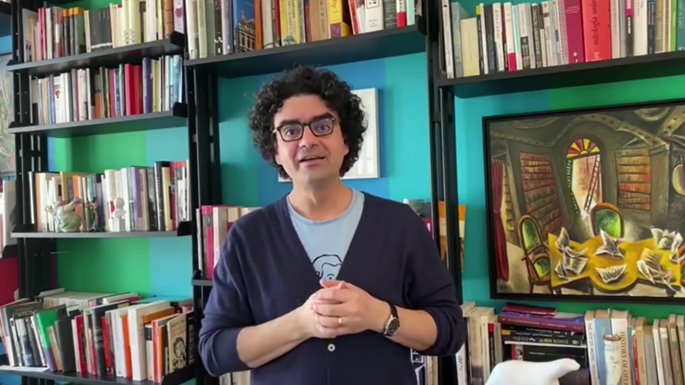 Stars von morgen@home: Rolando Villazón steht vor einer Wand mit Büchern und Bildern. Die Hände sind gefaltet. Er spricht in die Kamera.