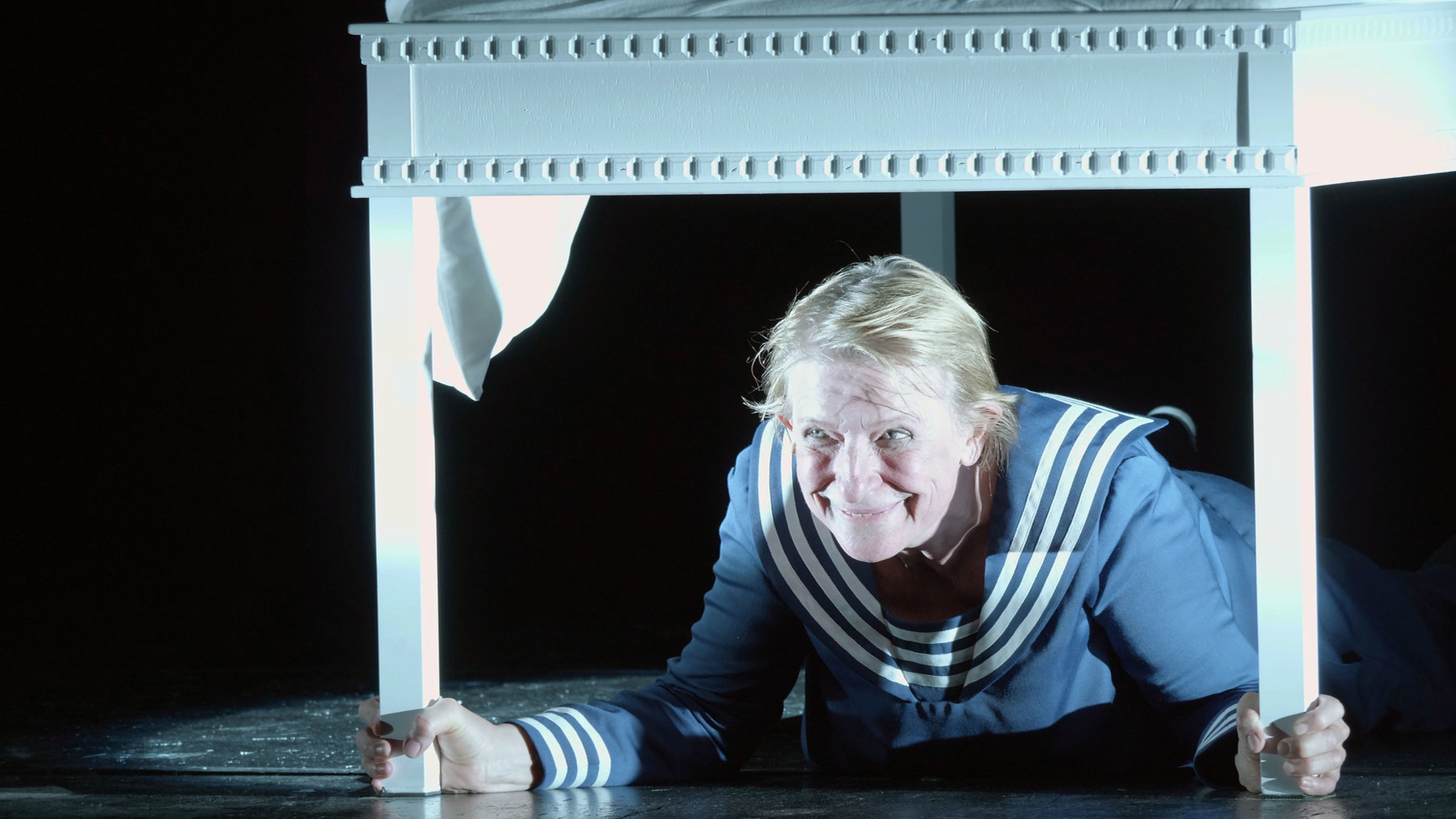 „THEATER.MACHER.INNEN“: Dagmar Manzel liegt in einem Matrosen-Kostüm und einem weißen Tisch und hält mit beiden Händen zwei Tischbeine in der Hand. Sie grinst verschmitzt nach vorne.