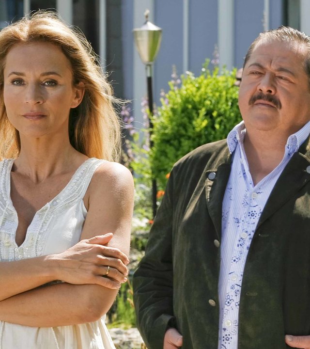 "Die Rosenheim-Cops - Mord auf Raten": Vor dem Treppenaufgang zur Terrasse eines Wohnhauses steht Cosima Tauber (Caroline Beil) mit verschränkten Armen im Garten und schaut in die Ferne. Hinter ihr steht Kommissar Hofer (Joseph Hannesschläger mit den Händen in den Jackentaschen und sieht sie an.