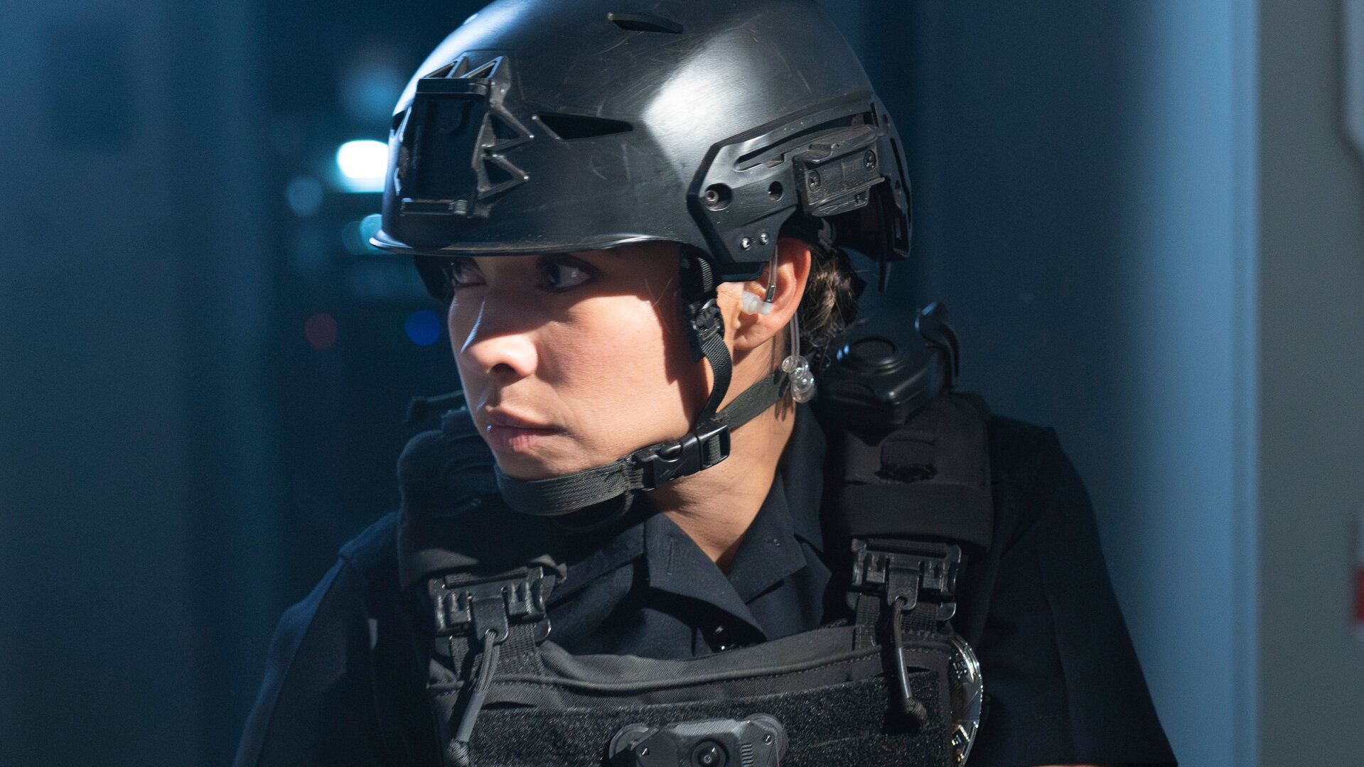 "The Rookie - April, April": Celina Juarez (Lisseth Chavez) steht in einem abgedunkelten Raum und trägt eine kugelsichere Weste sowie einen schwarzen Helm. In der Hand hält sie eine Pistole und blickt konzentriert nach vorn.