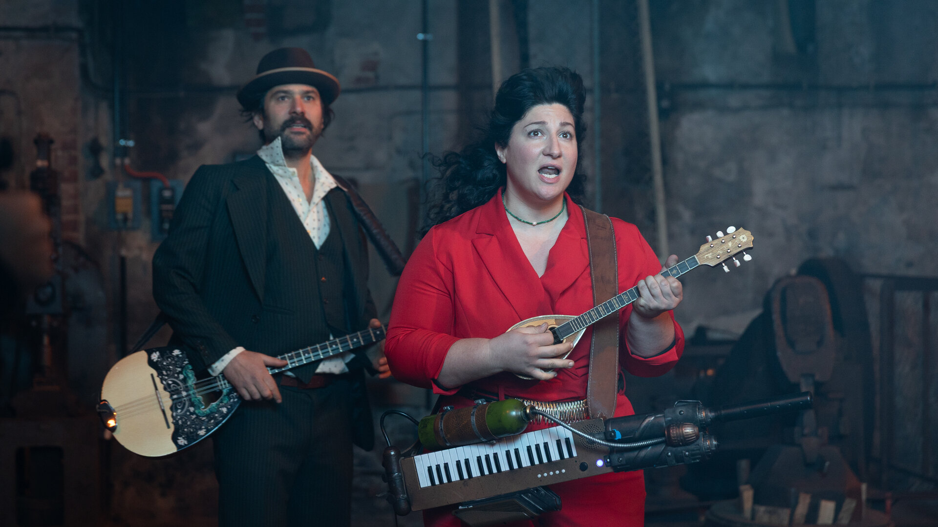"Rembetis - Die Geisterjäger - The Winner Takes It All": In einer alten Halle mit Maschinen im Hintergrund stehen Paris (Jasin Challah) und Alitheia (Fevronia). Alitheia spielt ein kleines Saiteninstrument und trägt ein selbstgebaute Melodica um die Hüfte. Daneben steht Paris in einem elegant Anzug mit Hut und hält eine Bouzouki. Beide wirken konzentriert.