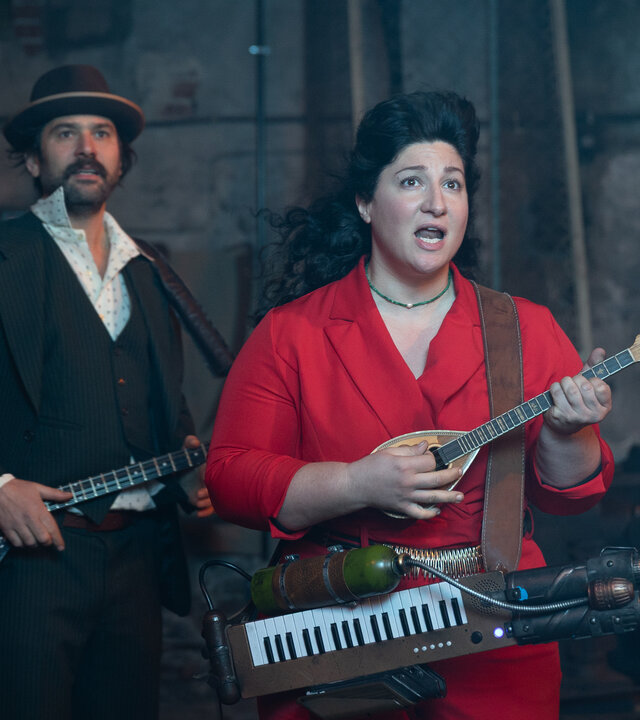 "Rembetis - Die Geisterjäger - The Winner Takes It All": In einer alten Halle mit Maschinen im Hintergrund stehen Paris (Jasin Challah) und Alitheia (Fevronia). Alitheia spielt ein kleines Saiteninstrument und trägt ein selbstgebaute Melodica um die Hüfte. Daneben steht Paris in einem elegant Anzug mit Hut und hält eine Bouzouki. Beide wirken konzentriert.